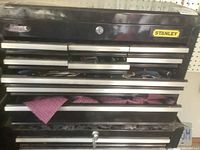 Front view of Stanley branded black metal tool box with multiple drawers containing hand tools and cloths; shows key lock but no key present.