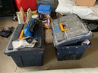 Two large dark blue plastic Mastercraft tool cases stacked in garage with tools and bags around them