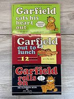 Three Garfield books stacked: 'Garfield Eats His Heart Out' (green cover), 'Garfield Out to Lunch' (red cover), 'Garfield Rolls On' (black cover).