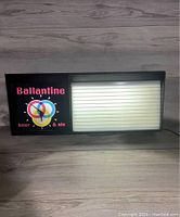 Front view of the lighted wall sign on a wooden floor with the light turned on, showing the Ballantine branding and letter panel.