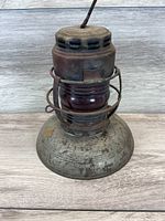 Front and side view of the Dietz Embury No. 40 lantern showing the red glass globe and the metal cage surrounding it. The lamp base shows rust and wear.