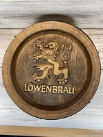Top view of wooden barrel lid featuring Löwenbräu lion logo and text carved in relief on wood surface.
