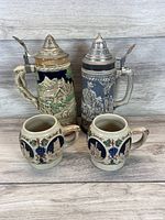 Two vintage beer steins with decorative designs and pewter lids, paired with two small ceramic mugs with scenic motifs and grapevine decoration.