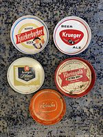 Five vintage round metal beer trays arranged on a carpet floor showing front graphics and wear.
