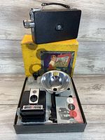 Photo showing Brownie movie camera in original box with yellow packaging, additional lens caps, and manual.