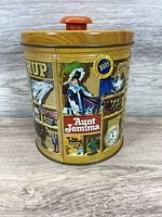 Front and side view of vintage Aunt Jemima pancake syrup tin showing detailed vintage graphics and wood-look lid with red knob.