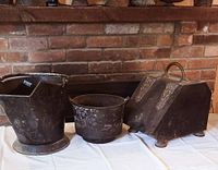 View of all three cast iron containers including a box coal scuttle and two ash buckets on a white cloth against brick backdrop
