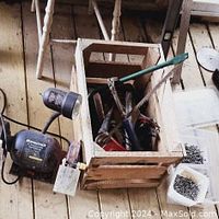 Wooden crate filled with vintage hand tools including wrenches, saws, and pliers, placed on wooden floor next to two boxes with nails and screws.