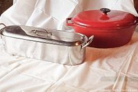 Wide shot showing the large red Dutch oven and the stainless steel fish steamer basket side by side on a white background.
