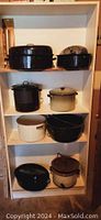 Full view of all pots on white shelving unit showing assortment of black, cream, white, and brown enamel pots and roasting pans with lids.