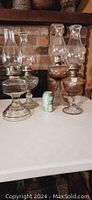 Front view showing all four hurricane oil lamps grouped together on a white table against a brick background. Two lamps have clear glass bases, two have bronze-tone metal bases. Each lamp has a clear glass chimney and visible wick mechanisms.