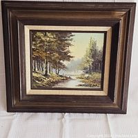 Framed oil painting on board showing forest and river landscape in 19th century style, with a wooden brown frame and signature visible on bottom right.