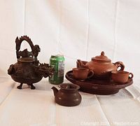 Cast metal dragon-themed teapot with intricate handle and spout next to small clay Gongfu teapot and plastic Gongfu tea set with teacups and tray with bird design