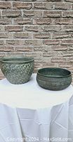 Two green glazed pots on a white cloth-covered table in front of a brick wall. The taller pot shows a scaled pattern, and the shorter pot displays intricate line work.