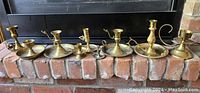 Seven vintage Victorian style brass candle and taper holders arranged in a row on a brick hearth in front of a fireplace, showing varying sizes and designs.