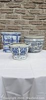 Four blue and white porcelain planters displayed on a white tablecloth against a brick background. Two round planters, one octagonal planter, and one two-piece flower pot with stand visible.