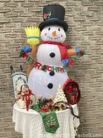 Inflatable snowman with black hat and broom holding colorful revolving disco light in torso, displayed outdoors on table with various Christmas decorations around.