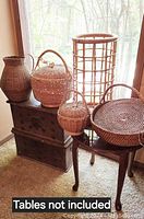 Six assorted handwoven baskets displayed on wooden tables and chests next to a window for lighting. Showing their shapes, sizes, and styles.