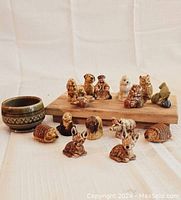 Image showing collection of 17 various small ceramic figurines of animals and human characters arranged on and around a wooden board, alongside a small brown ceramic bowl.