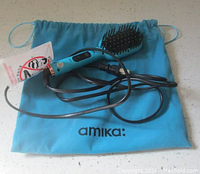 Mini straightening brush placed on top of blue velvet drawstring pouch with 'amika:' branding. The brush has a light blue handle, black ceramic bristles with black nylon cool-tips, and attached power cord.
