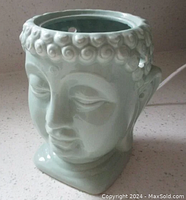 Front-left angled view of light green ceramic Buddha head lamp on countertop with power cord visible.