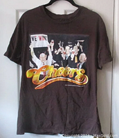 Front view of dark brown t-shirt with Cheers TV show characters graphic and 'Cheers' logo on front