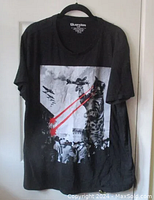Full front view of black cotton t-shirt on hanger featuring large graphic of giant cat with red laser eyes shooting planes and crowd.