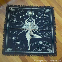 Top-down photo of black altar cloth with central golden goddess figure, sun and moon symbols, starry border, and fringed edges on wooden floor.