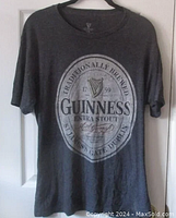 Full front view showing the dark gray T-shirt with the Guinness Extra Stout label printed in white and black with a gold harp detail.