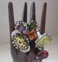 Front view of six rings displayed on a wooden ring holder; large animal print ring with rhinestones, multi-colored earring with purple center, rectangular yellow stone, orange square stone, red teardrop stone, and green teardrop stone