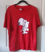 Front view of red T-shirt on a hanger showing full Snoopy letter graphic and floating heart detail.