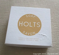 Top view of white box with gold circular logo reading HOLTS SOAP SAVON, with text 'Vanilla Oat and Honey' in both English and French on the front. Box lid shows slight tear on front.