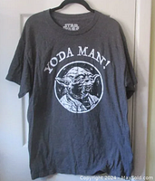 Gray large size T-shirt with white front graphic of Yoda and 'Yoda Man!' text, hanging on plastic hanger against white door.