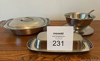 Three stainless steel kitchen serving items: covered vegetable dish, butter dish, gravy boat with ladle.