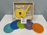 Front angle view of the Fisher Price record player showing the cream plastic body, yellow turntable and tonearm, and 5 colorful records arranged in front.