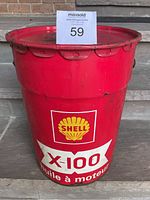 Red large cylindrical vintage metal oil can with Shell logo, X-100 branding, and French text 'huile à moteur'. Hinged lid with rim indentations, paint wear and chipping visible.