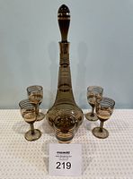 Front view of the MCM decanter set on a white patterned cloth, showing decanter with stopper and five glasses arranged around it, highlighting the gold detailing on amber-colored glass.
