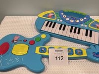 Top-down view of the Peppa Pig blue toy guitar and keyboard on a white cloth background showing details of buttons and keys with the lot number 112 tag.