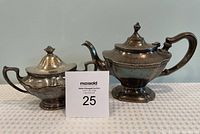 Silver-plated tea pot and sugar bowl placed side by side on a white surface with auction lot number card between them