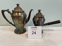 Two silver-plated tea pots with lids on a white textured surface. The left pot is tall with a shield emblem and ornate handle; the right pot is shorter with a detachable spout and straight handle.