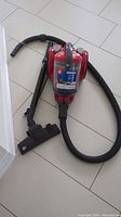 Red Dirt Devil Breeze vacuum cleaner with hose and black floor nozzle attachment shown on white tiled floor.