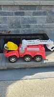 Full side view of the red toy firetruck with yellow plastic helmet on the rear seat, against a stone wall outside.