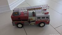 Front and side view of red and white firetruck toy with extendable ladder, decals and controls on side panel, wheels visible