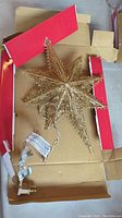 Gold Christmas tree star tree topper laid out on a piece of cardboard with original box sides visible. Shows wired string lights and power plug attached.