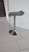 Gray stool with curved seat, adjustable height mechanism, chrome base and footrest on a tiled floor against a cream wall.