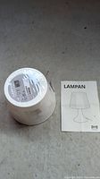 White IKEA Lampan lamp shade wrapped in plastic with IKEA label on bottom, alongside a folded instruction manual for the Lampan table lamp.