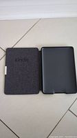Black Amazon Kindle Paperwhite and accompanying black protective case with textured finish and Kindle tab, both lying open on a tiled floor.
