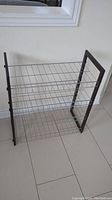 Front and side view of a modern metal shoe rack with three wire mesh shelves and black metal frame, standing on tiled floor against a white wall.