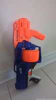 Side view of the orange and blue Nerf SurgeFire Elite blaster sitting on the floor, angle shows rotating drum and grip.
