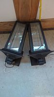 Two black cast iron outdoor lantern lights with glass panels shown from the front on concrete floor.